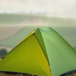 Gunung Nganten di Lencoh Selo, Camping Tanpa Trekking Berlatar Merapi Ilustrasi tenda hijau di lereng berumput saat fajar dengan kabut tipis di atas ladang