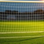 Ilustrasi gawang sepak bola kosong dengan jaring putih di lapangan rumput saat senja